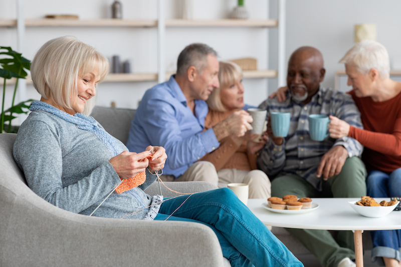 Relaxed senior woman knitting and sitting in arm chair by her friends drinking tea with cookies together, having a conversation and laughing