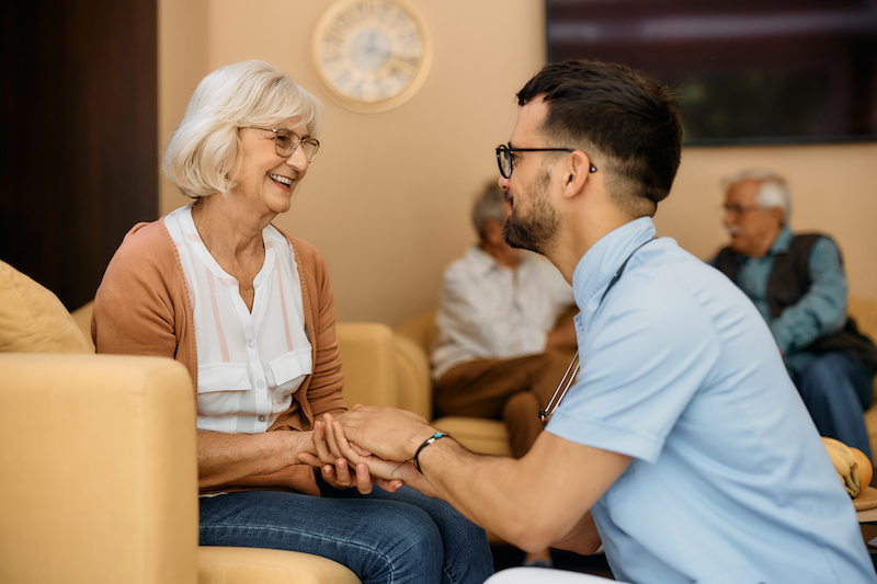 Happy male caregiver and senior woman hold hands while communicating at residential care home