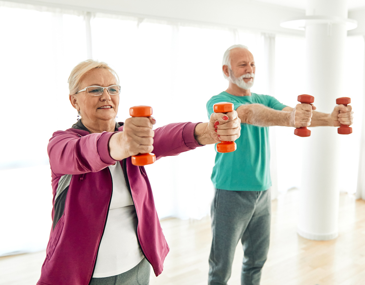 A middle-aged woman, standing further forward, and an elderly man, in the background, holding up a pair of dumbells in each hand. Depicts weight training routines for seniors